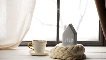 Das Foto zeigt eine dampfende Teetasse und ein Keramik-Haus vor einem Fenster.