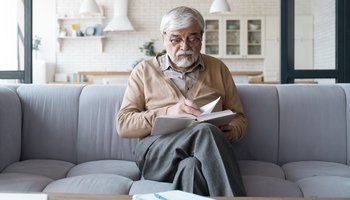 Ein älterer Herr mit grauen Haaren und Brille sitzt konzentriert lesend auf einem Sofa, ein Buch in der Hand haltend, in einem hellen, gemütlich wirkenden Raum.