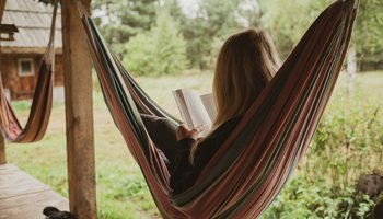Eine Frau liegt in einer Hängematte, die an einer Holzterrasse zwischen zwei Balken befestigt ist. Sie liest ein Buch und ist von hinten fotografiert, sodass ihr Gesicht nicht sichtbar ist.